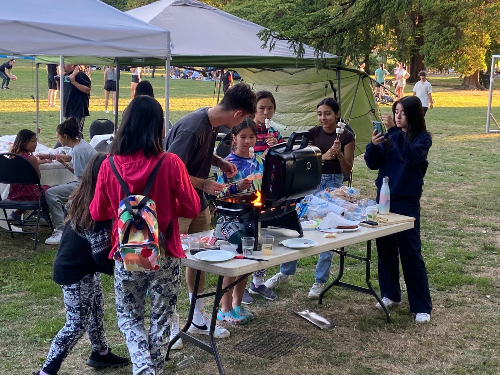 Junior youth and families gathered around a barbecue at a community park evening.